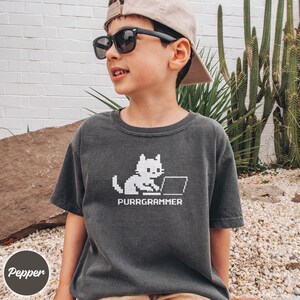 May include: A charcoal gray short-sleeve t-shirt featuring a white pixelated graphic of a cat sitting at a laptop computer. Below the cat, the text reads PURRGRAMMER in a matching white pixelated font style on the front of the garment.