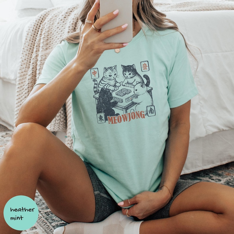May include: A mint green t-shirt featuring a graphic illustration of four cats playing mahjong around a table. The design includes the text MEOWJONG in orange letters and various Chinese characters in small squares surrounding the cat illustration.