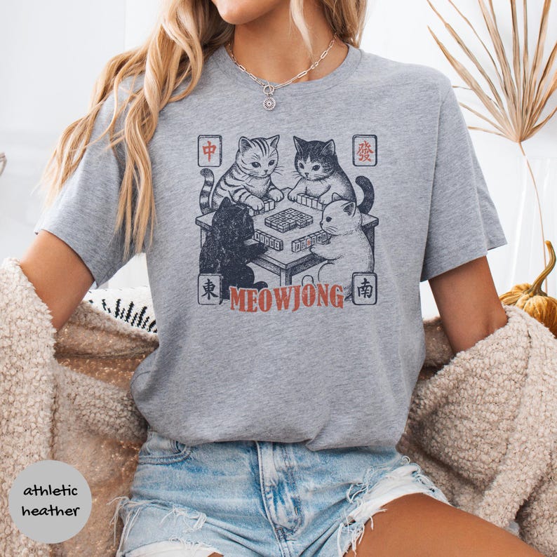 May include: An athletic heather gray t-shirt featuring a graphic of four cats playing mahjong around a table. The design includes Chinese characters in the corners and the text MEOWJONG in orange letters below the table. The shirt has short sleeves.