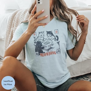 May include: A light blue t-shirt featuring a graphic print of four cats playing mahjong around a table. The design includes the text MEOWJONG in orange and various Chinese characters in small square frames surrounding the illustration of the cats.
