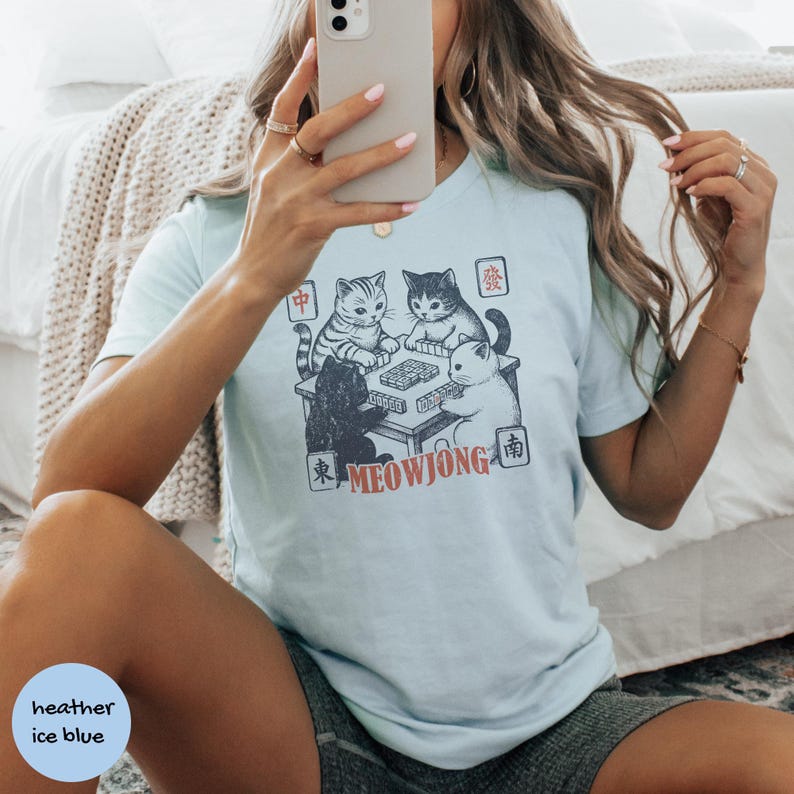 May include: A light blue t-shirt featuring a graphic print of four cats playing mahjong around a table. The design includes the text MEOWJONG in orange and various Chinese characters in small square frames surrounding the illustration of the cats.