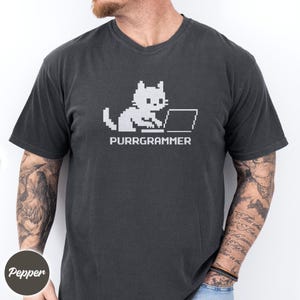 May include: A charcoal gray short-sleeve t-shirt featuring a white pixel art graphic of a cat sitting at a laptop computer. Below the cat, the text reads PURRGRAMMER in a matching white pixelated font style.