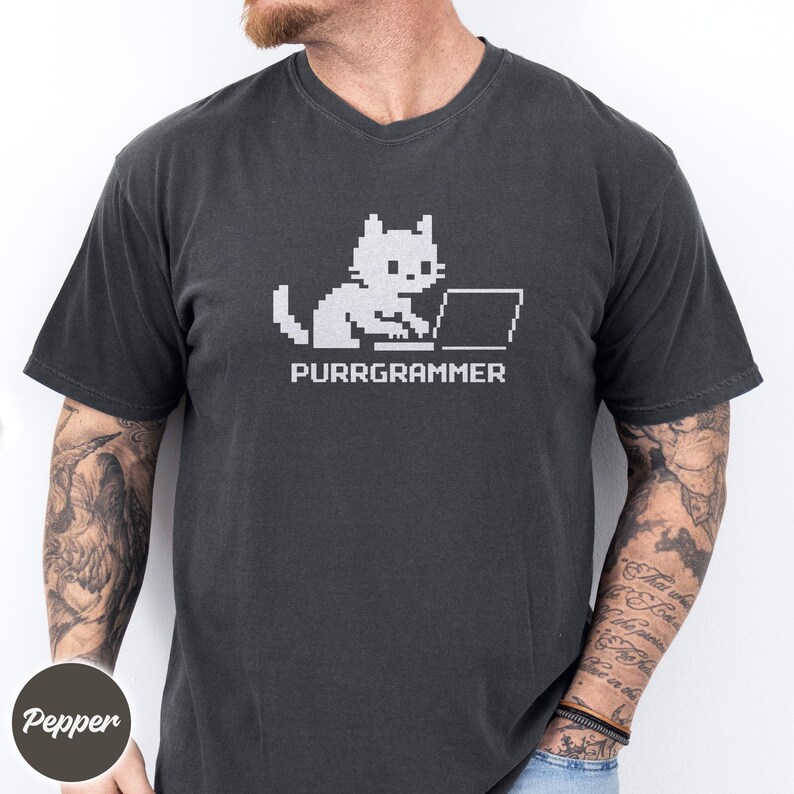 May include: A charcoal gray short-sleeve t-shirt featuring a white pixel art graphic of a cat sitting at a laptop computer. Below the cat, the text reads PURRGRAMMER in a matching white pixelated font style.