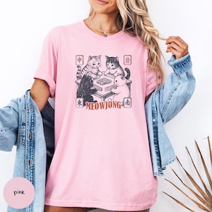May include: A light pink short-sleeved t-shirt featuring a graphic of four cats playing mahjong around a table. The design includes Chinese characters in four corners and the text MEOWJONG in a reddish-brown font below the table illustration.