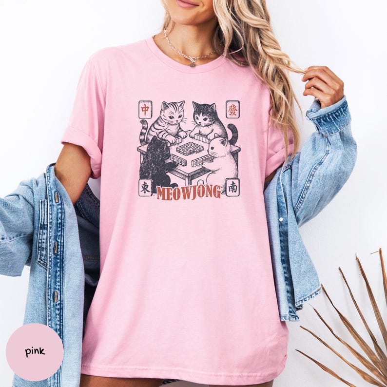 May include: A light pink short-sleeved t-shirt featuring a graphic of four cats playing mahjong around a table. The design includes Chinese characters in four corners and the text MEOWJONG in a reddish-brown font below the table illustration.