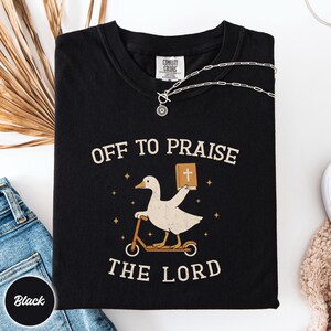 May include: A black short-sleeve t-shirt featuring a graphic of a white goose riding a scooter and holding a brown book with a white cross. The text "OFF TO PRAISE THE LORD" is printed in a beige font above and below the goose illustration.