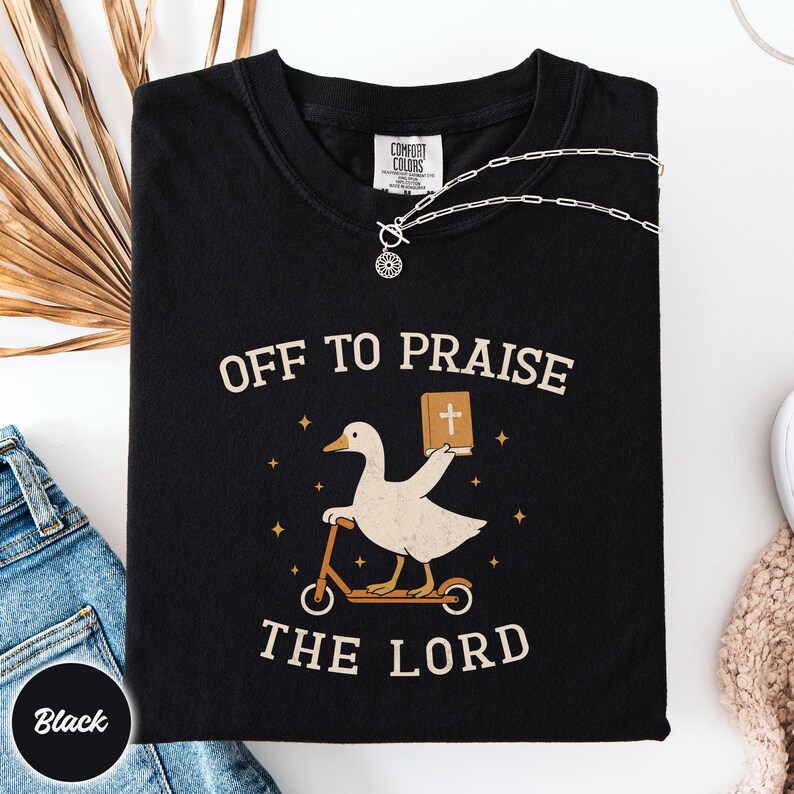 May include: A black short-sleeve t-shirt featuring a graphic of a white goose riding a scooter and holding a brown book with a white cross. The text "OFF TO PRAISE THE LORD" is printed in a beige font above and below the goose illustration.