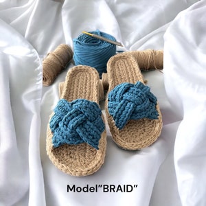 May include: A pair of handmade crochet slippers with a braided blue top detail. The slippers are beige with a blue accent, and the text "Model 'BRAID'" is visible at the bottom. The slippers are displayed on a white surface.