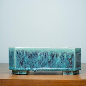 Op de afbeelding: Een rechthoekige keramische plantenbak met een turquoise en donkerblauwe glazuur. De plantenbak heeft een laag profiel, met een reeks kleine voetjes. Het glazuur heeft een gevlekt uiterlijk, met donkerdere kleurvlakken. De plantenbak staat op een houten oppervlak.