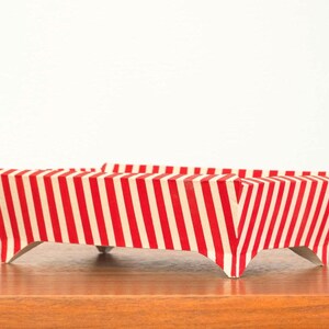 May include: A rectangular tray with a red and white striped pattern. The tray is elevated on three small, silver-colored legs. The tray is sitting on a wooden surface.