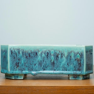 May include: Rectangular turquoise ceramic planter with a textured surface and dark brown accents. The planter has a low profile, raised on four small feet. A decorative piece for plants.