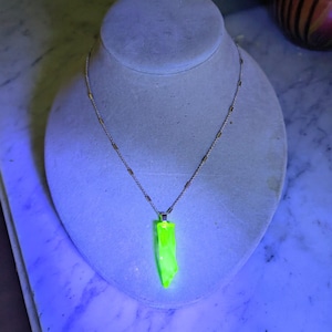 May include: A silver-toned chain necklace featuring a long, pointed pendant that glows with a bright, neon-green fluorescent light. The pendant has a smooth, translucent appearance and is suspended from a delicate metal chain with small, evenly spaced cylindrical links.