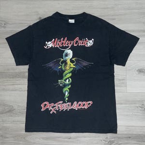 May include: Black t-shirt with the band name "Mötley Crüe" in white and red script above a graphic of a staff with wings and a green snake. The text "Dr. Feelgood" is below the graphic in red.