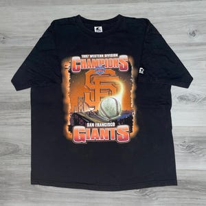 May include: Black t-shirt with a graphic commemorating the 1997 Western Division Champions, San Francisco Giants. The design includes the team's logo, a baseball, and the Golden Gate Bridge, with orange and white accents. The Starter logo is visible on the sleeve.