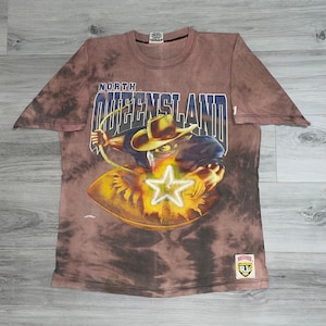 May include: Brown tie-dye t-shirt with a graphic featuring a cowboy roping a star, with the text "NORTH QUEENSLAND" in blue and white. The shirt has a crew neck and short sleeves.