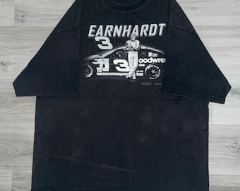 Puede incluir: Camiseta negra con el nombre "EARNHARDT" sobre un gráfico de un coche de carreras y un piloto, con el número "3" en blanco. La camiseta tiene cuello redondo y mangas cortas. El diseño es en blanco y gris.