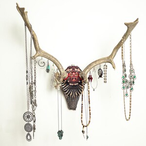 May include: A wall-mounted deer skull with large antlers used as a jewellery organiser. The skull features a black and white striped pattern with red accents at the top. Various silver and gold-toned necklaces and earrings hang from the antlers.
