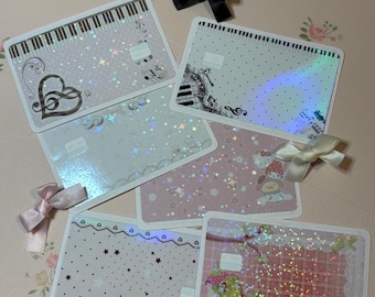 May include: A collection of decorative cards with musical and character-themed designs. The cards feature piano keys, musical notes, and star patterns, with a pink and white color scheme. Small bows in black and pink add a decorative touch.