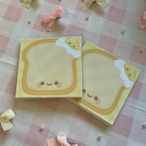 May include: Two square notepads with a cute cartoon design of toast and a fried egg. The toast is light yellow with a brown outline, and the egg has a white and yellow design. The notepads are on a pink and white checkered surface.