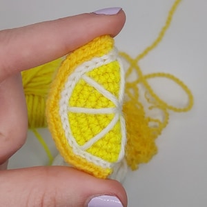 May include: A crocheted lemon slice with a white centre and yellow outer edges. The lemon is held in a hand.