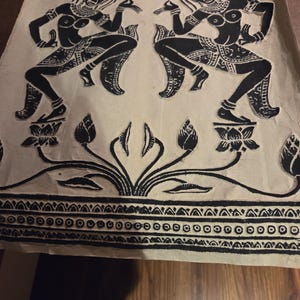 Könnte beinhalten: Ein rechteckiges Blatt cremefarbenes Papier mit einem schwarzen Blockdruck. Das Motiv zeigt zwei gespiegelte Figuren in Tanzposen, die jeweils auf einer Lotusblüte stehen, über einer dekorativen Bordüre mit geometrischen Mustern.