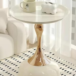 May include: A round white side table with a glossy finish, featuring a metallic gold pedestal base and a wide, rounded white bottom. On the tabletop sits a cream-coloured ceramic mug with a small illustration and a white, cloud-shaped decorative object.
