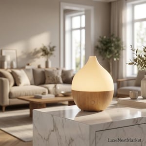 May include: A teardrop-shaped essential oil diffuser with a light-coloured wood grain base and a smooth, matte white upper section that emits a soft, warm glow. A small circular power button is centred on the front of the wooden base.