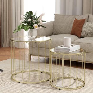 May include: A set of two round nesting tables featuring clear glass tops and gold-toned metal frames with vertical wire detailing. The tables have a cylindrical structure with open sides and small black feet at the base for stability on the floor.