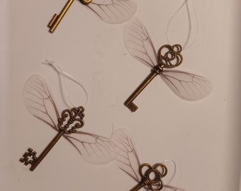 May include: Four antique-style keys with delicate, translucent wings. The keys are made of gold-toned metal and have intricate designs. The wings are a light, almost transparent, white.