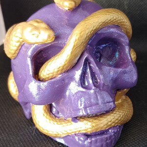 May include: Purple skull with gold snakes wrapped around it. The snakes are textured and have a shiny finish. The skull has a iridescent finish.