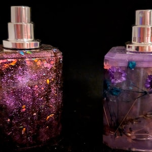Puede incluir: Dos botellas de vidrio con tapas de pulverización plateadas. La botella de la izquierda está llena de un líquido morado brillante y la botella de la derecha está llena de un líquido transparente con flores secas.