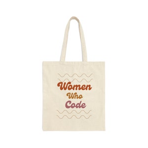 Peut inclure: Sac fourre-tout crème avec le texte "Women Who Code" dans une police rétro. Le texte est dans des tons de marron et de violet, avec des lignes ondulées au-dessus et en dessous. Le sac a de longues anses.