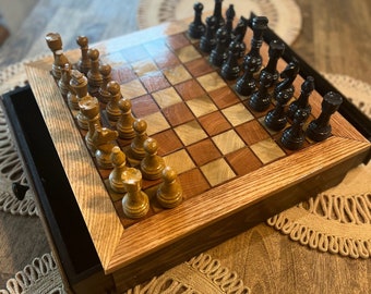 May include: A wooden chessboard with a drawer for storing the pieces. The board is made of light and dark wood and has a checkered pattern. The pieces are black and white and are arranged in a starting position. The board is sitting on a table with a brown surface.