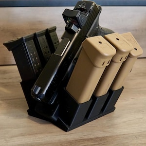 Puede incluir: Una pistola negra con mira de punto rojo se muestra en un soporte negro, junto con varios cargadores. El soporte tiene ranuras para el arma y los cargadores, con tres cargadores color canela y tres negros.