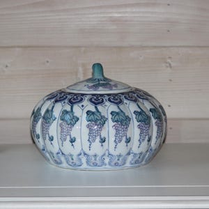 May include: A white ceramic lidded jar with a blue and purple grape design. The jar has a round shape and a decorative lid.