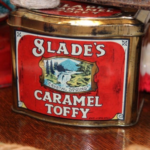 May include: A vintage tin box with a red label and gold trim. The label reads "Slade's Crystal Spring Caramel Toffee" with a small image of a waterfall and trees. The box is slightly worn and has a rustic look.