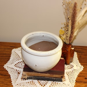May include: A white ceramic pot filled with brown sand sits on top of a stack of books. The pot is on a white crocheted doily. A small glass vase with dried flowers sits on the table next to the pot.
