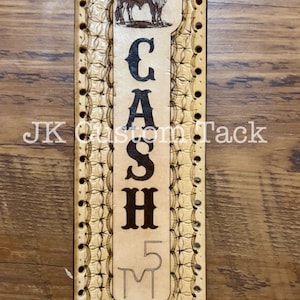 May include: A tan leather belt strap with a decorative pattern and the word "CASH" in black lettering. The strap also features a small image of a cow and the number "5" with the letter "M" below it.