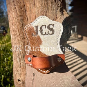 May include: A brown and white cowhide leather belt hanger with a brown leather strap and silver metal accents. The hanger is personalized with the initials "JCS".