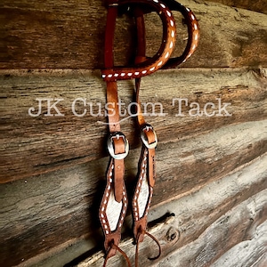 May include: A pair of brown leather stirrup leathers with white stitching and a white hair-on hide insert. The leathers are hanging on a wooden wall with the text "JK Custom Tack" visible.