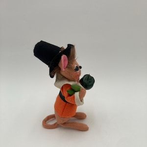 May include: A plush mouse figurine dressed in a pilgrim outfit. The mouse wears an orange jacket, a black hat, and a white collar. It holds a green object, possibly a pumpkin, in its paws. The figurine has pink ears and a long tail.