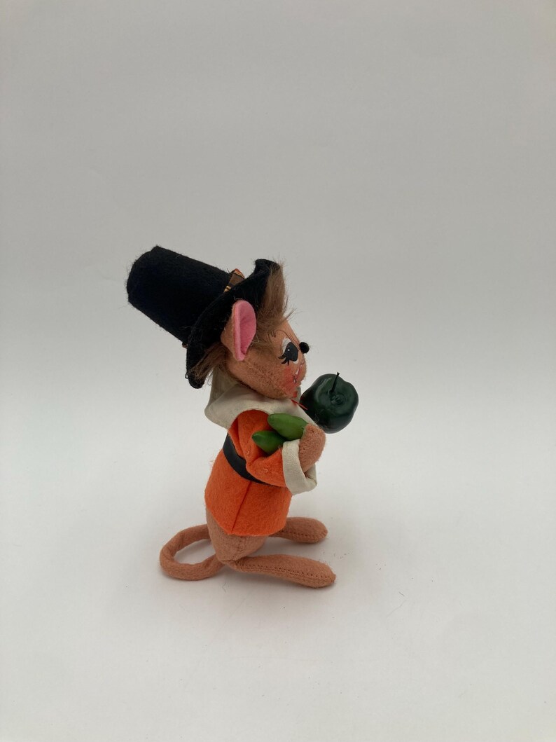 May include: A plush mouse figurine dressed in a pilgrim outfit. The mouse wears an orange jacket, a black hat, and a white collar. It holds a green object, possibly a pumpkin, in its paws. The figurine has pink ears and a long tail.