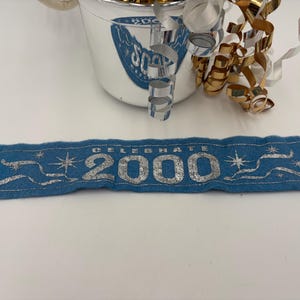 May include: A blue felt banner with the text "CELEBRATE 2000" in silver lettering. The banner has silver star and ribbon accents. A silver and white bucket with "2000" text and gold and silver curling ribbon are in the background.