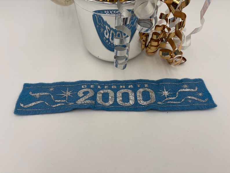 May include: A blue felt banner with the text "CELEBRATE 2000" in silver lettering. The banner has silver star and ribbon accents. A silver and white bucket with "2000" text and gold and silver curling ribbon are in the background.