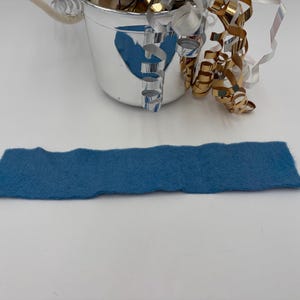 May include: A rectangular piece of blue felt with a wavy edge is displayed. A small silver bucket with a handle is in the background, filled with shiny gold and silver decorative ribbons. The felt is approximately 10 inches long.
