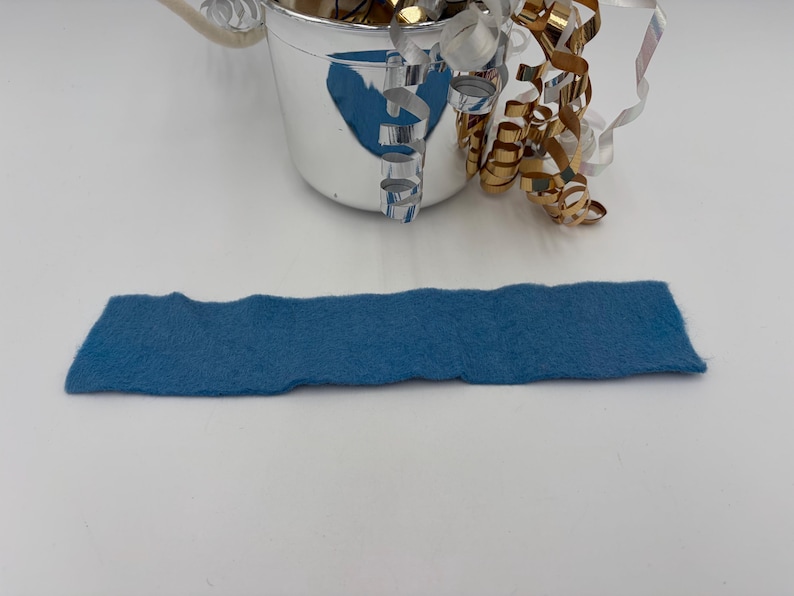 May include: A rectangular piece of blue felt with a wavy edge is displayed. A small silver bucket with a handle is in the background, filled with shiny gold and silver decorative ribbons. The felt is approximately 10 inches long.