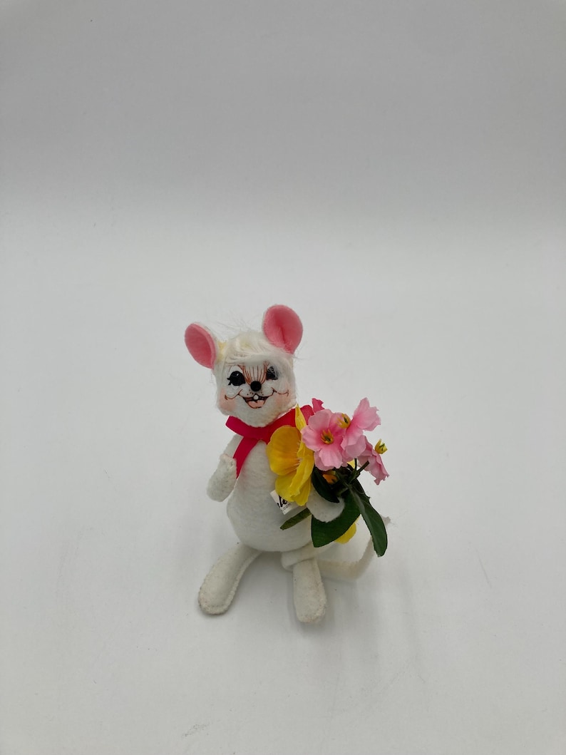 K&ouml;nnte beinhalten: Eine wei&szlig;e Filzmaus mit rosa Ohren und einer roten Schleife h&auml;lt einen Strau&szlig; gelber und rosa Blumen.