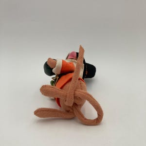 May include: A handmade felt doll in a seated position, wearing an orange vest, black hat, and white-trimmed sleeves. The doll has a pink face and brown limbs. A small green and gold charm is attached to the vest.