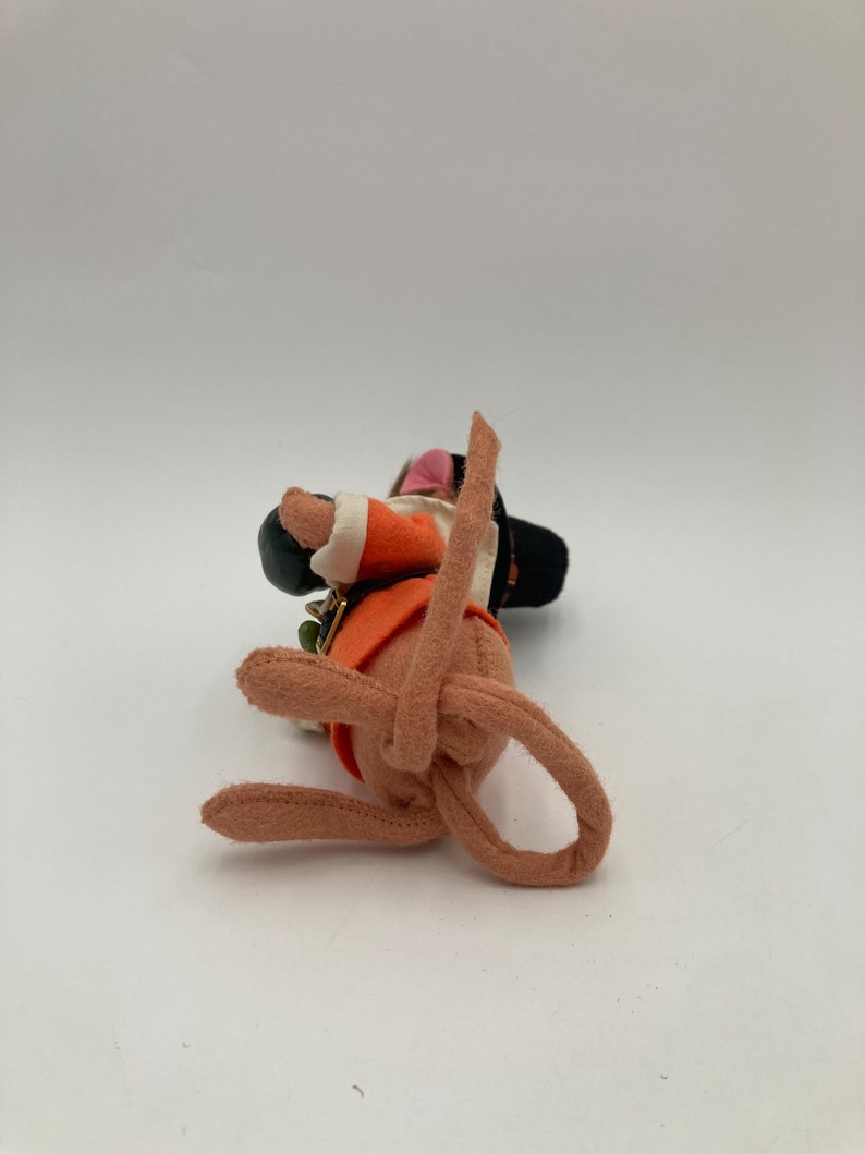 May include: A handmade felt doll in a seated position, wearing an orange vest, black hat, and white-trimmed sleeves. The doll has a pink face and brown limbs. A small green and gold charm is attached to the vest.