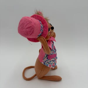 May include: A brown plush mouse wearing a pink top hat, a floral vest, and a pink bow. The mouse is holding the hat, with its ears and whiskers visible. The toy is set against a white background.